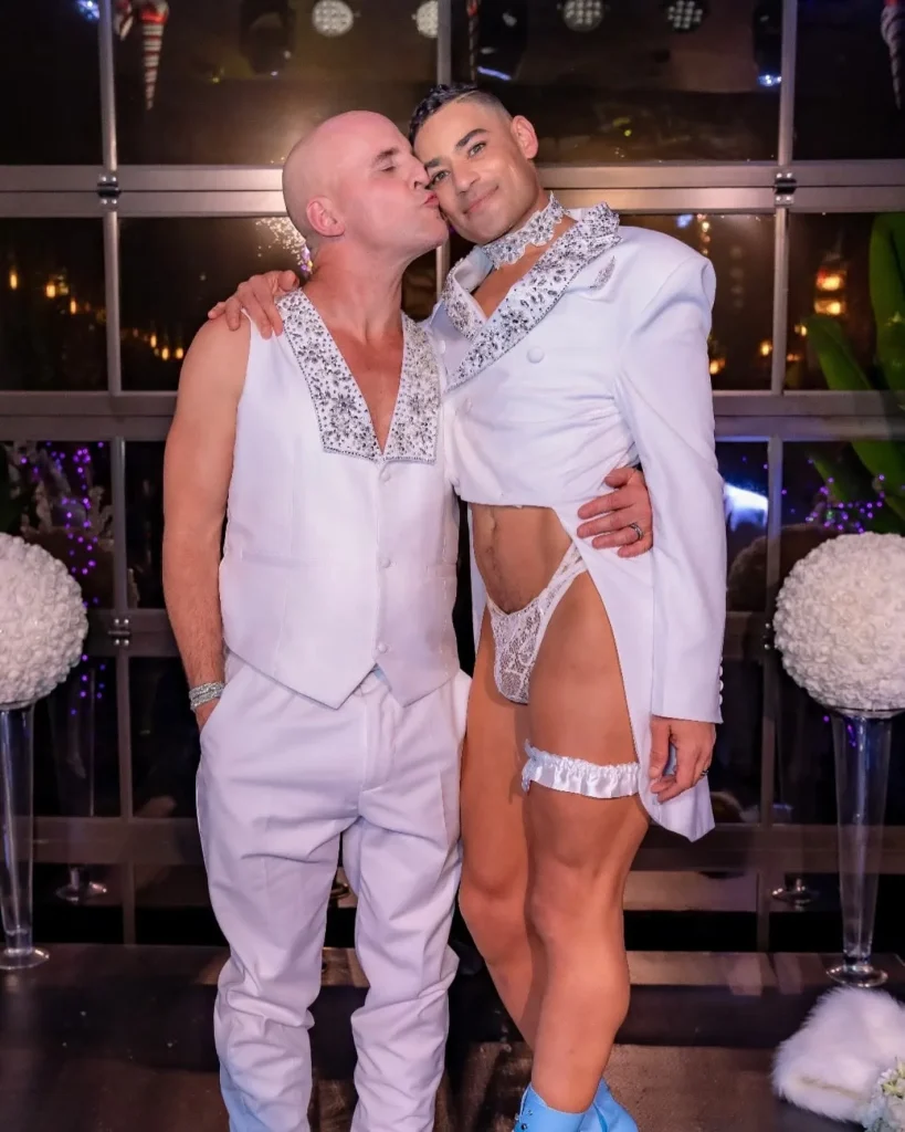 Two people in coordinated white formalwear, one kissing the other on the cheek